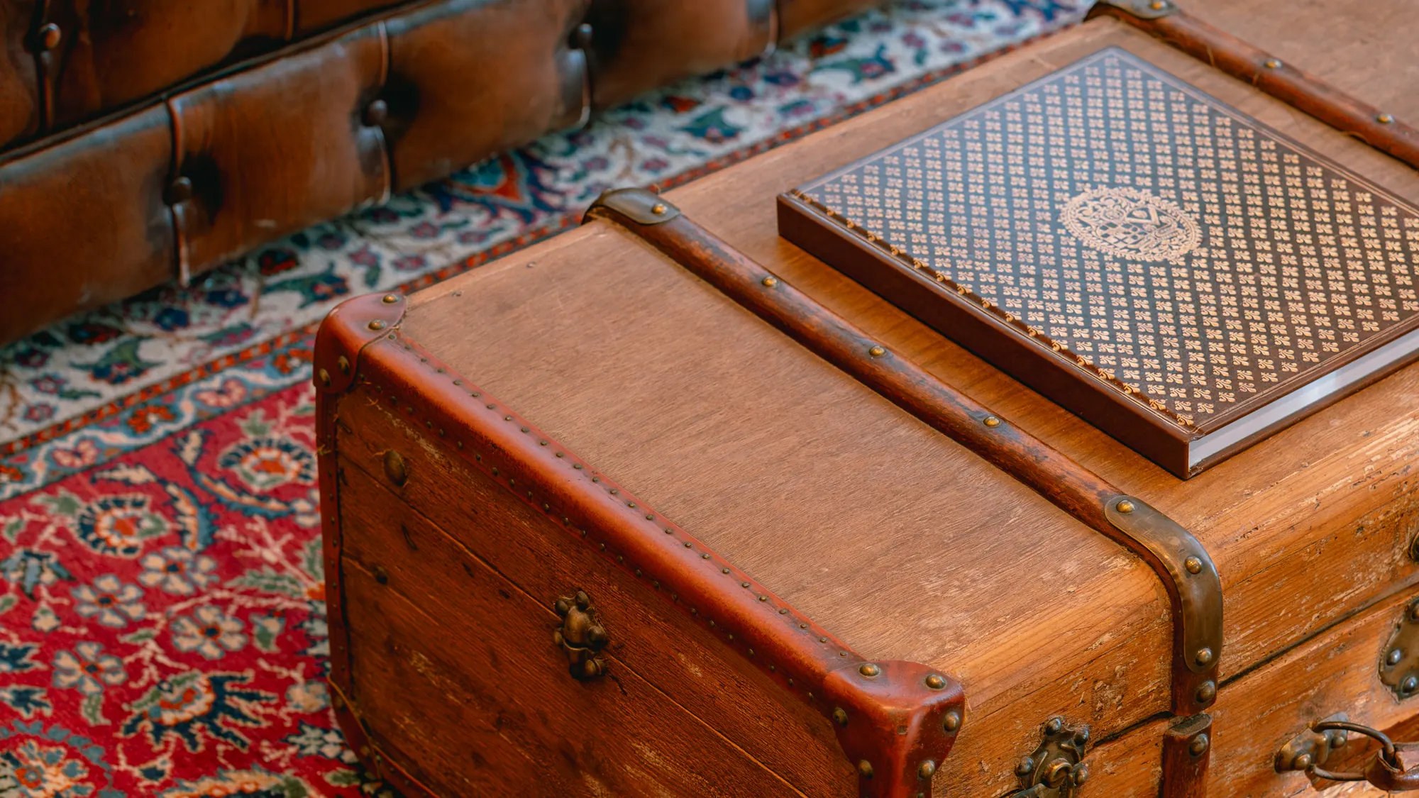 Un salon : une malle de voyage en bois posée sur un tapis, un livre relié dessus, une banquette de cuir derrière.